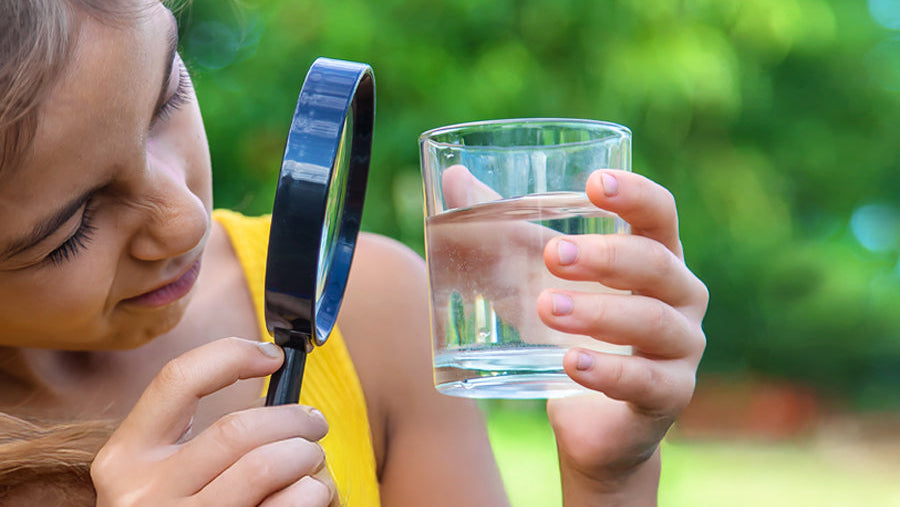 girl in yellow dress holding a glass of water and a magnifying glass outdoors.